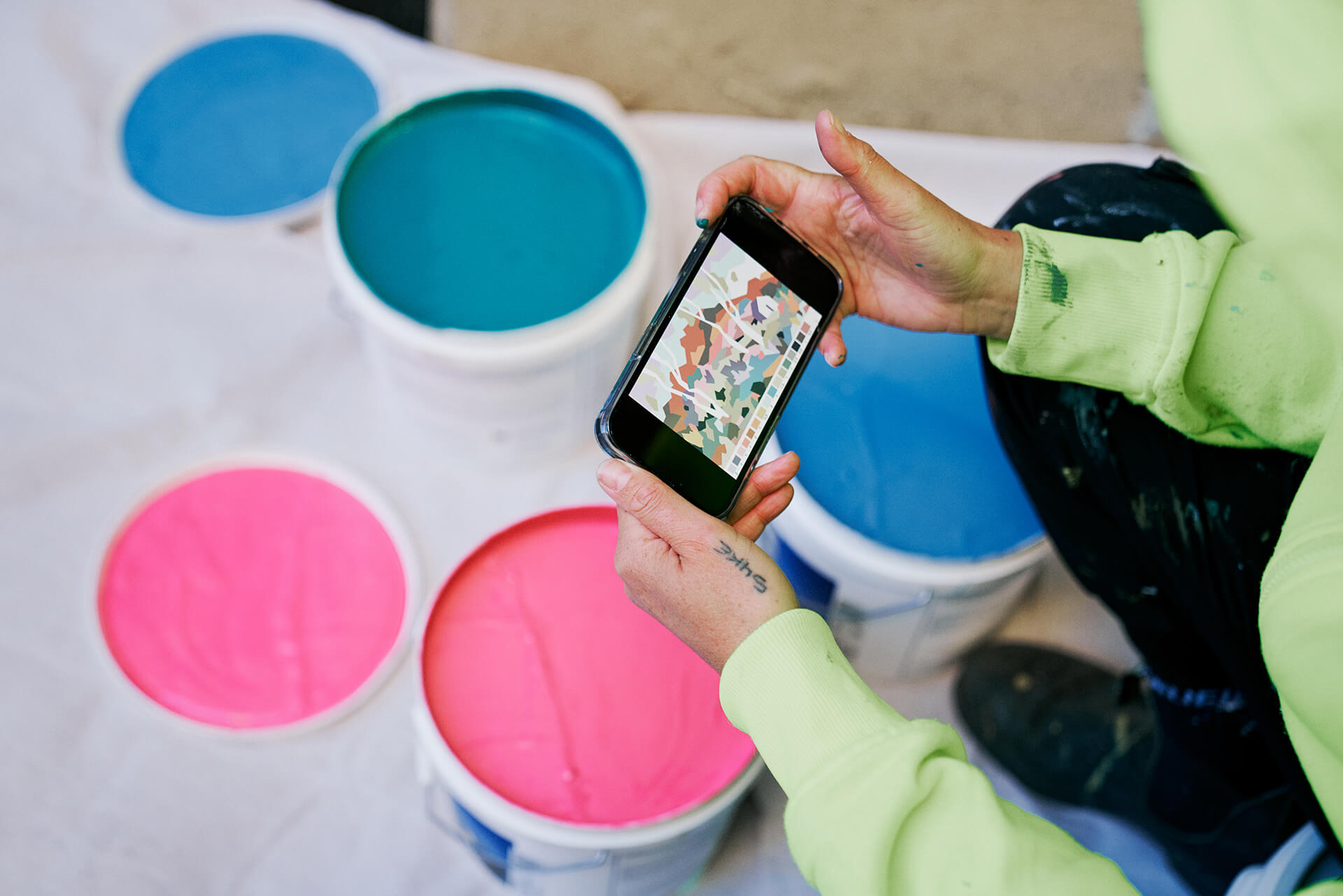 Artist holding a phone over tins of bright paint featuring a reference image of a geometric multicoloured wall mural produced by VANDAL for Mirvac